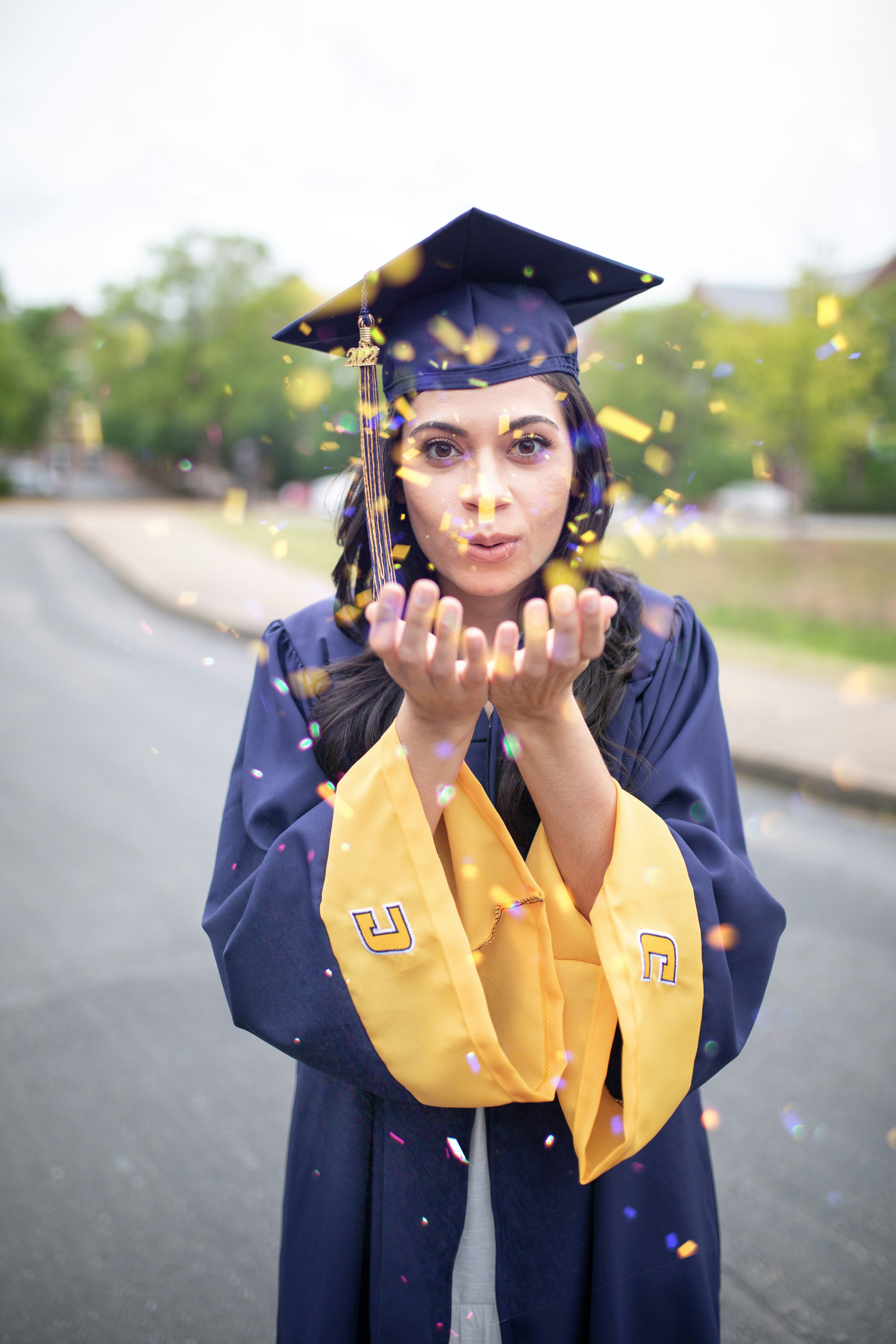 latina blowing yellow glitter into the camera. she is wearing a blue and yellow graduation cap and gown